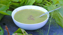 Une assiette creuse contenant de la soupe verte et une cuillere est installée sur une ardoise avec des légumes verts autour